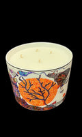 A three-wick scented candle in a white ceramic pot with a colorful, textured graphic design featuring a prominent orange leaf motif, set against a dark gray textured fabric background.