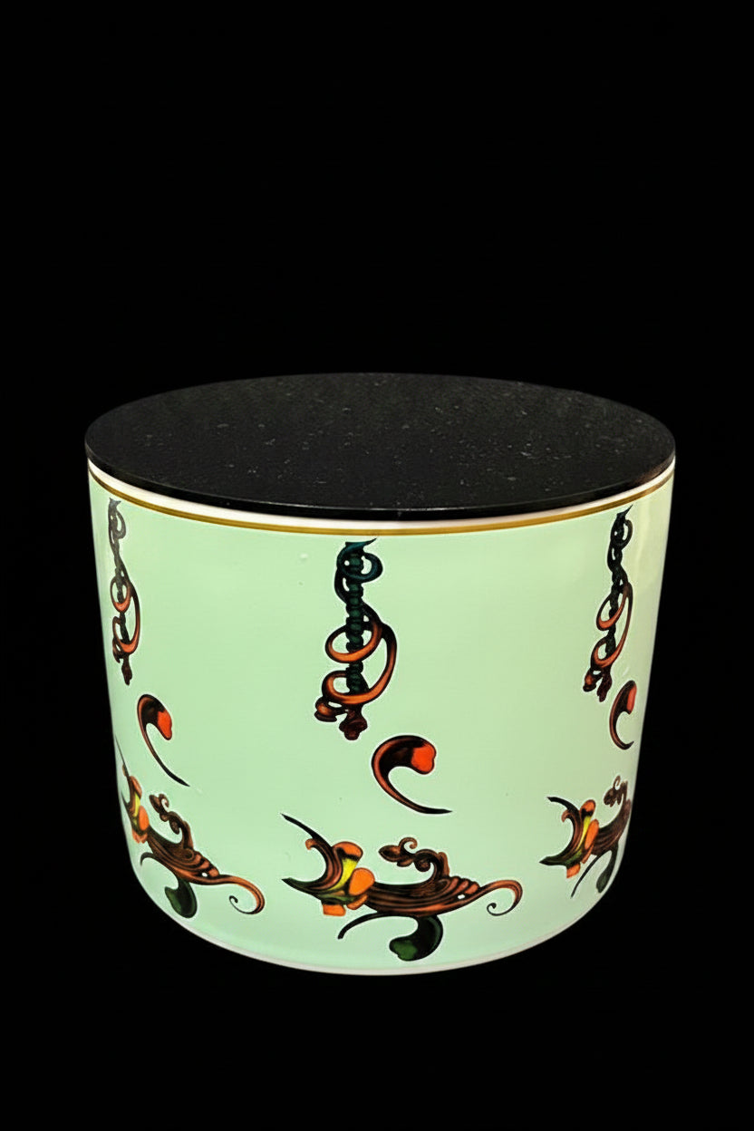 A cylindrical ceramic container with a pale mint green body decorated with black, orange, and gold abstract scrollwork, featuring a solid black lid, set against a dark gray textured fabric background.