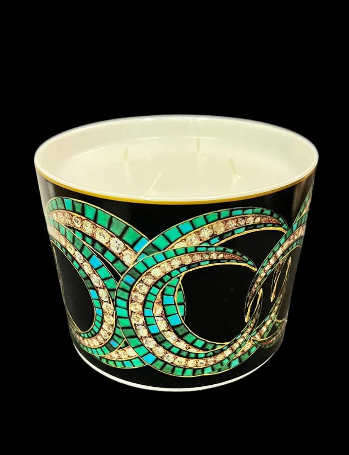 A black ceramic candle vessel with a white wax interior and a decorative Art Deco style pattern featuring turquoise green, gold, and black swirls, set against a gray textured fabric background.
