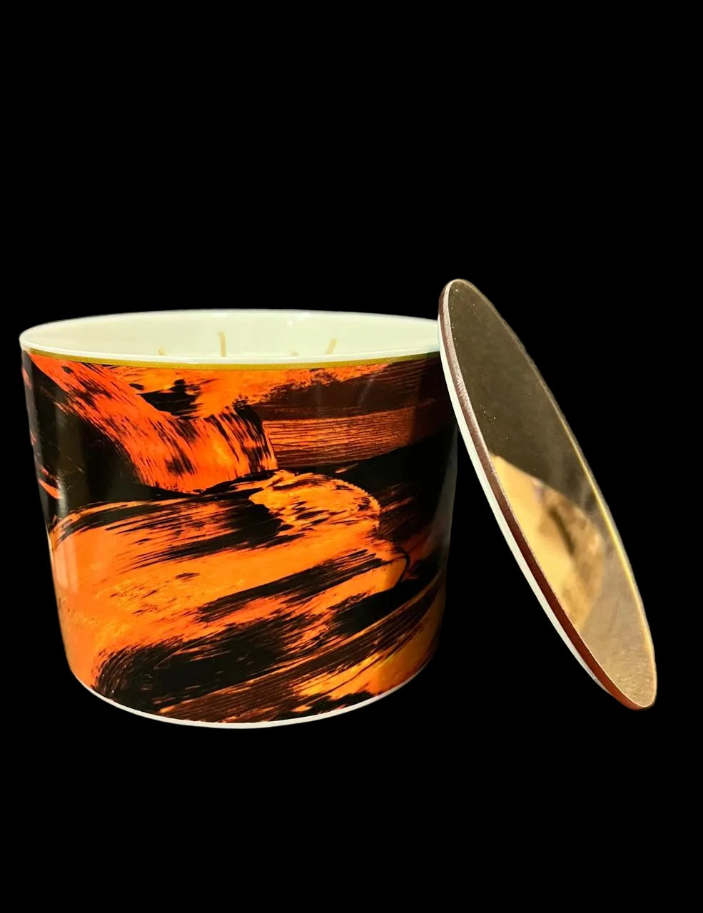 A cylindrical candle tin with a dramatic orange and black landscape-style print, shown with its lid leaning against it on a textured gray fabric background.