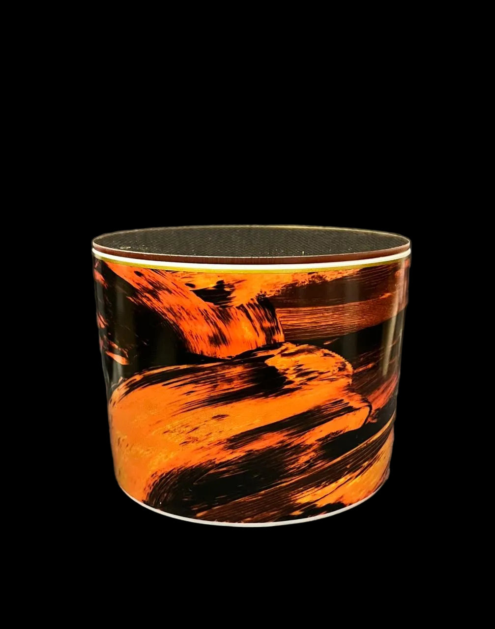 A cylindrical container or cup with a glossy black and orange abstract brushstroke pattern, photographed against a dark gray textured fabric background.