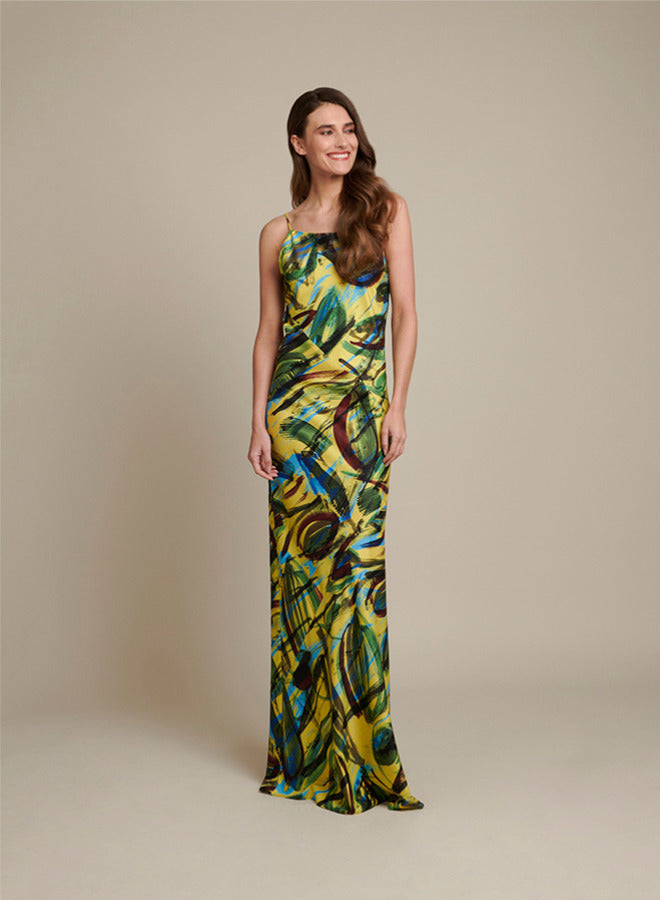 A woman modeling a sleeveless, floor-length maxi dress with a vibrant abstract print in yellow, green, blue, and brown tones, set against a solid beige studio background.