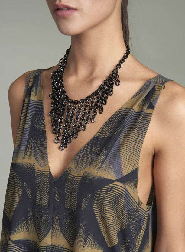 A close-up of a model wearing a multi-strand black beaded necklace over a sleeveless V-neck top with a blue, gray, and gold abstract pattern.