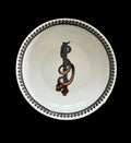 A white ceramic bowl with a black beaded rim and a central dark brown abstract spiral motif, placed on a square matching plate featuring a bold black, gray, white, and yellow graphic pattern.