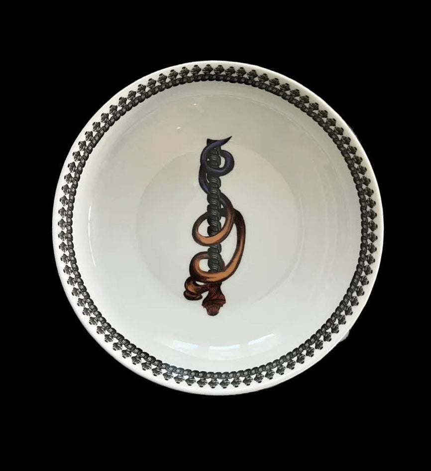 A white ceramic bowl with a black beaded rim and a central dark brown abstract spiral motif, placed on a square matching plate featuring a bold black, gray, white, and yellow graphic pattern.