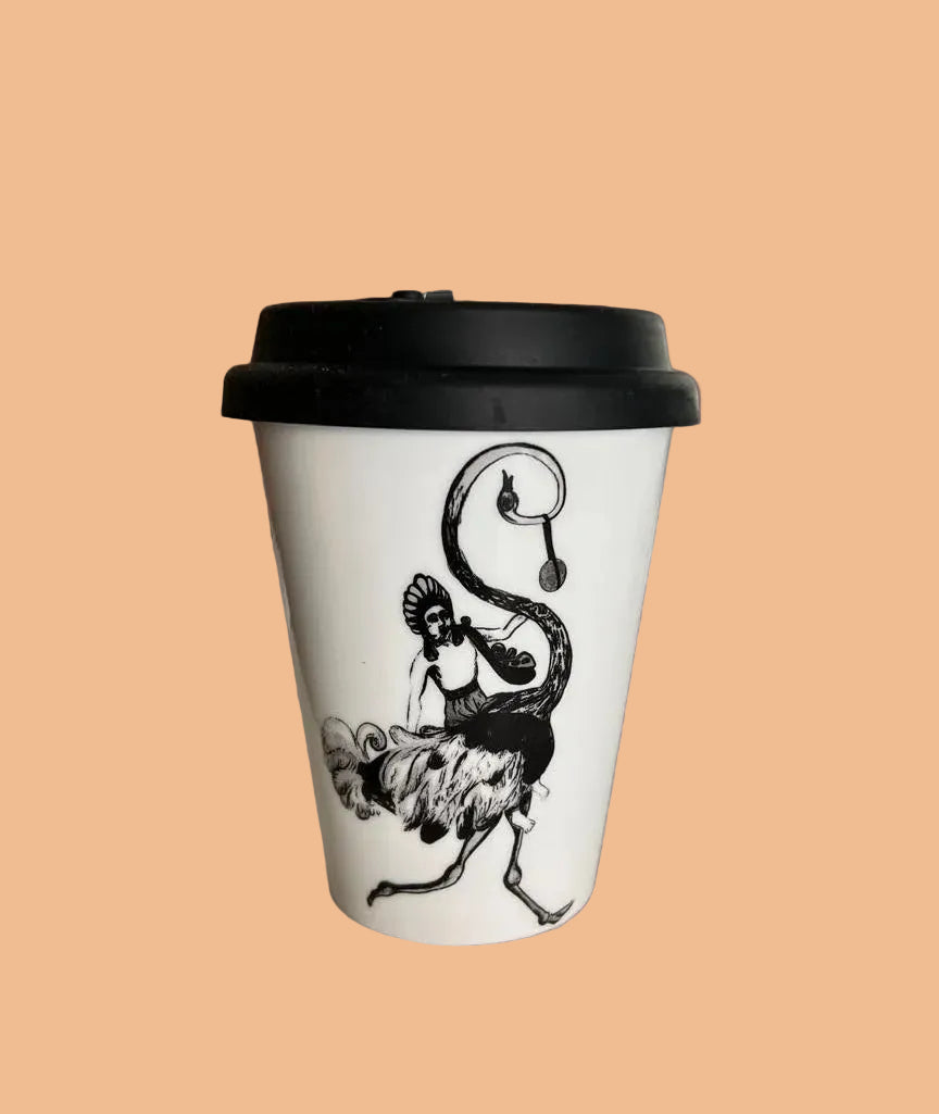 A white paper coffee cup with a black lid, featuring a black line art illustration of a stylized figure, sitting on a light-colored tabletop in a blurred indoor setting.
